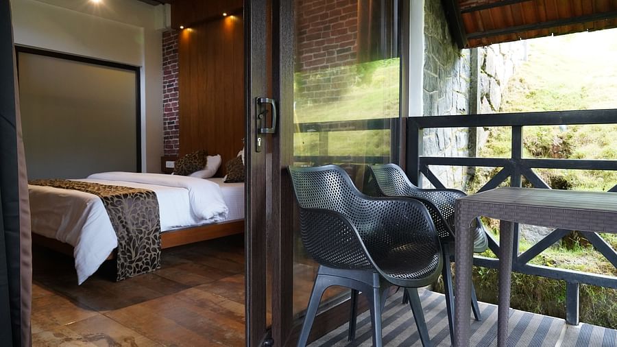 A bed reflected in a glass door, with a small desk and chair near the balcony. | Premium Forest View Cottage With Balcony | The Wild Trails Munnar