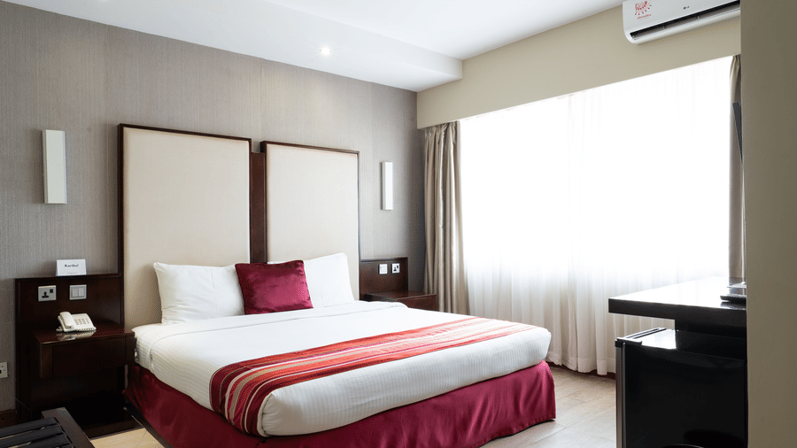 A hotel room at The Zehneria, Nairobi, featuring a double bed, an AC unit, a writing table, and night stands on either sides of the bed.