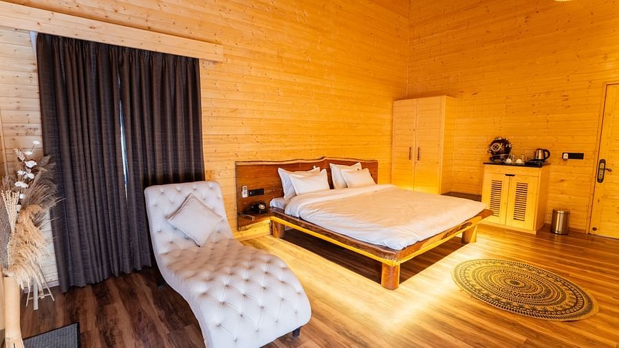 A warmly lit wooden cabin-style room at Themis Farm House features a double bed, a patterned chaise lounge, dark curtains, and subtle under-bed lighting.