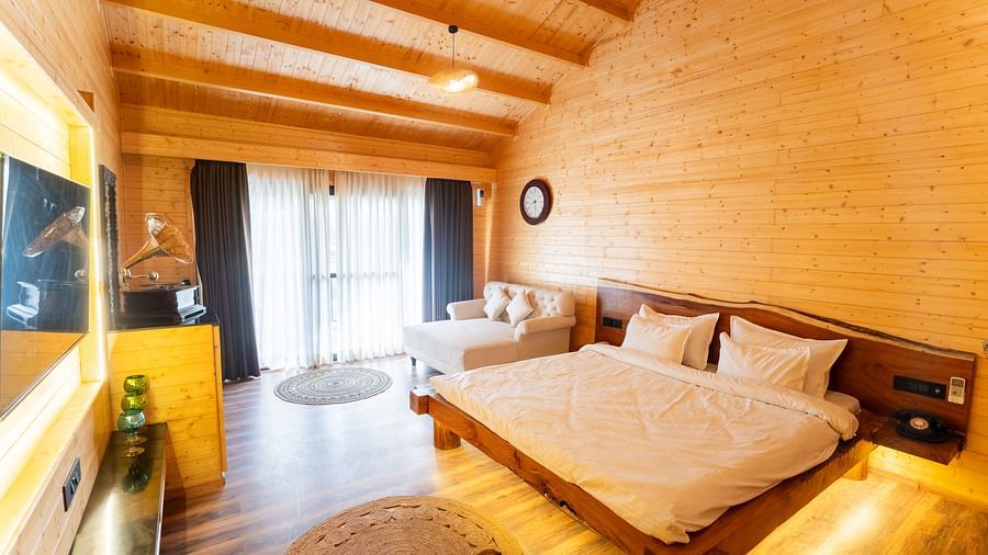 A warmly lit room at Themis Farm House features wooden walls and ceiling, a double bed with white linens, a seating area by a balcony, and a wall-mounted TV.