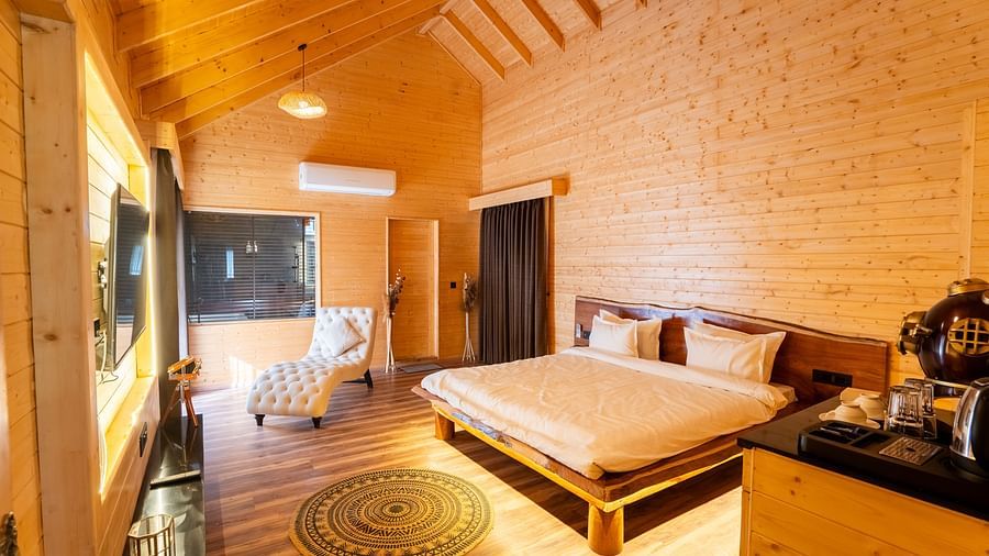 A warmly lit hotel room at Themis Farm House features wooden walls and a high ceiling, a double bed, a patterned chaise lounge, and a television.