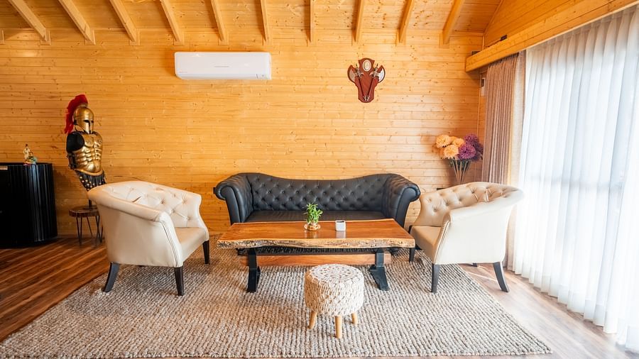 A well-lit lounge area with wooden walls and ceiling features a black leather sofa, two cream armchairs, a wooden coffee table, a small stool, and decorative items including a knight figurine at Themis Farm House.