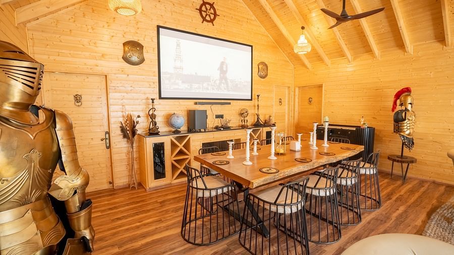 A room with wooden walls and a high ceiling features a long dining table with stools, decorative items including armour figures, and a large screen at Themis Farm House.