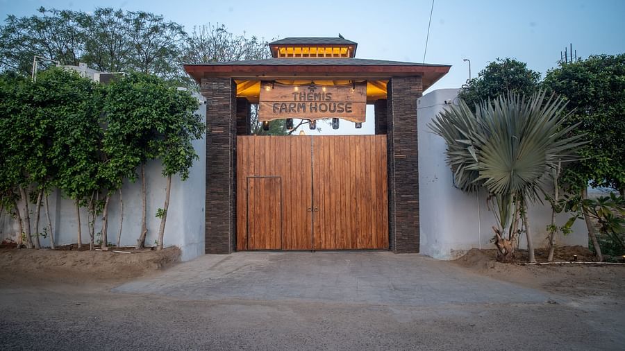 Grand wooden entrance gate of Themis Farm House, flanked by lush greenery and palm trees, set against a serene, blue-sky backdrop.