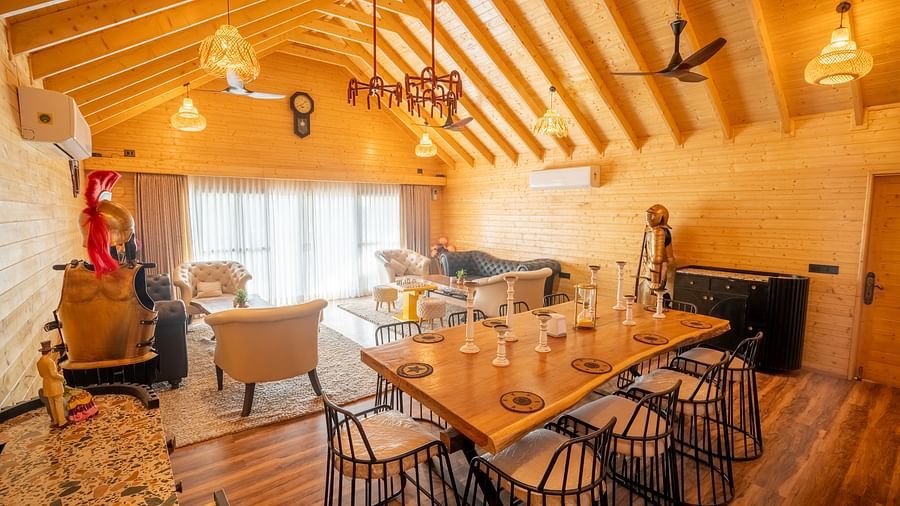 A spacious room with a high wooden ceiling and various light fixtures features a long wooden table with chairs, comfortable lounge seating, and decorative items at Themis Farm House.