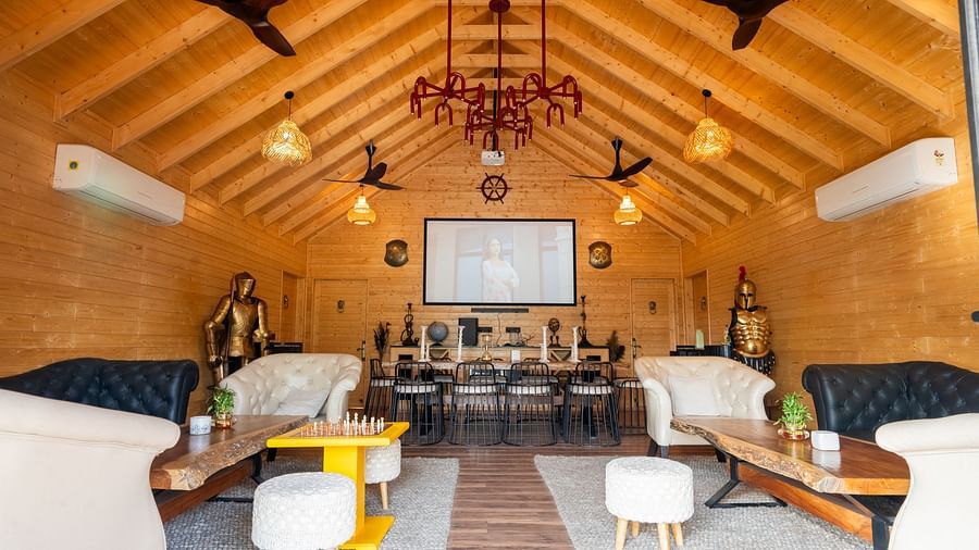 A spacious room with a high wooden ceiling and multiple fans features lounge seating with sofas and stools, alongside a dining area with tables and chairs at Themis Farm House.