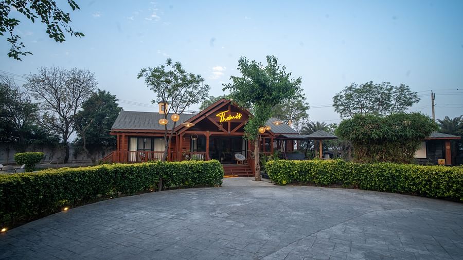Front view of Themis Farm House with warm lighting, wooden exterior, lush greenery, and a spacious driveway under a clear evening sky.