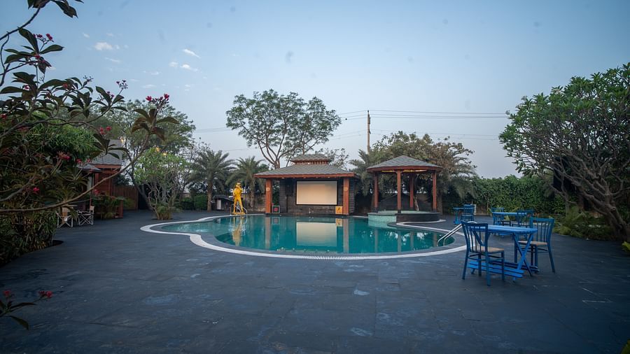A tranquil swimming pool at Themis Farm House surrounded by lush greenery, relaxation pavilions, and blue outdoor furniture under a clear evening sky