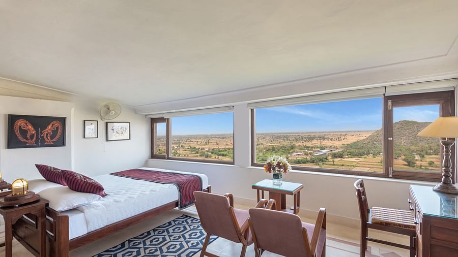 A room with a daybed, a set of chairs, a small table, a desk, framed artworks, and large windows with a scenic view in Tijara Fort-Palace - 19th Century, Alwar.