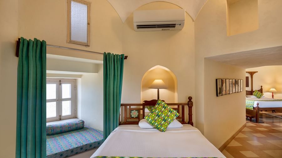 Room with bed, patterned runner and pillow, curtains, window seat, arched alcove with lamp, wall-mounted air conditioner, and doorway leading to another room - Tijara Fort-Palace - 19th Century, Alwar.