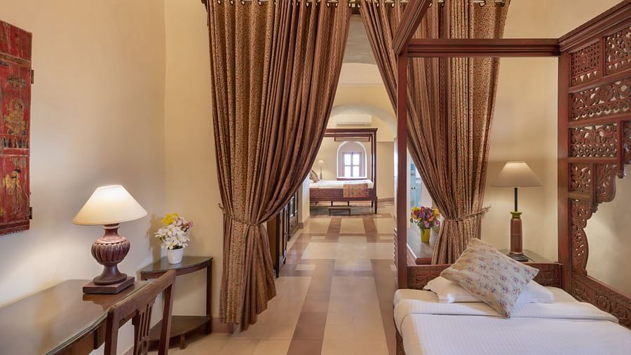 Room with four-poster bed, patterned pillow, lamp, table, curtains leading to another room with visible bed, and patterned floor - Tijara Fort-Palace - 19th Century, Alwar.