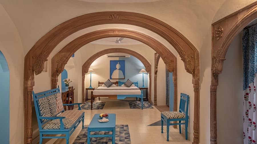 A hotel suite with arched doorways, a seating area with a blue sofa and coffee table, a blue chair, and a view into a bedroom with a bed and bedside tables with lamps at Tijara Fort-Palace - 19th Century, Alwar.