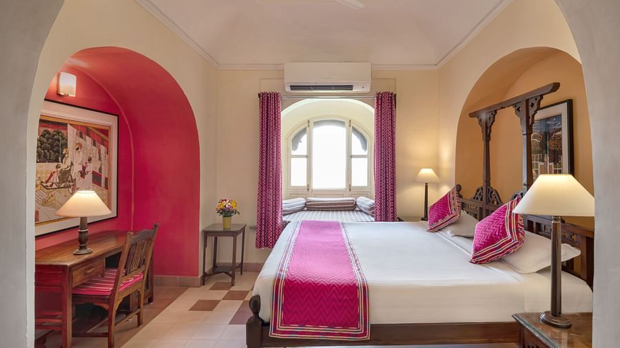 Hotel room with a four-poster bed featuring pink accents, bedside tables with lamps, a window seat with purple curtains, a desk in a red alcove, and AC at Tijara Fort-Palace, Alwar.