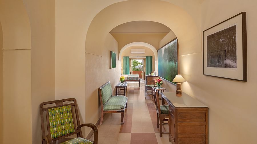 Hotel hallway with arched openings, a chair, a bench, a desk with a lamp, artwork, and a view into a room with a bed and seating at Tijara Fort-Palace, Alwar.