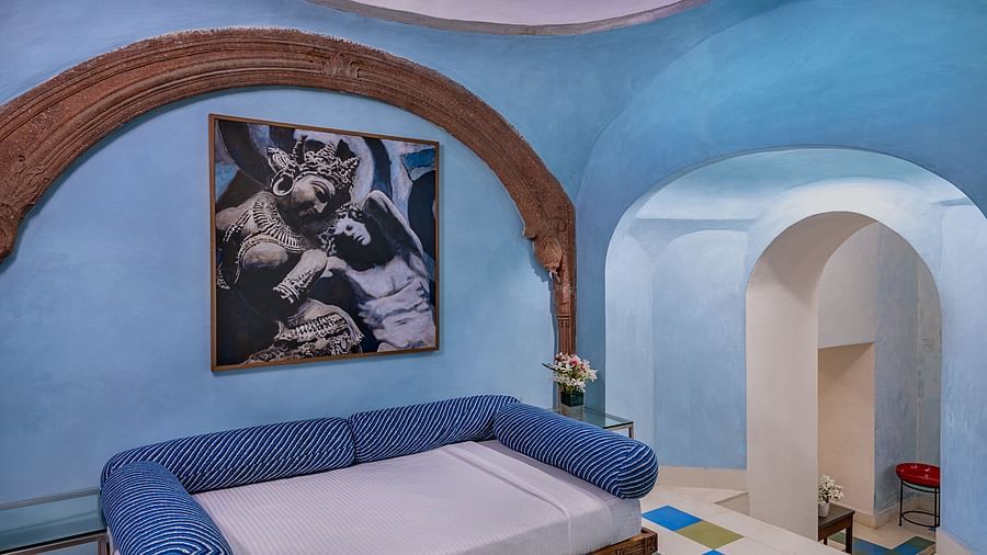 A room with a daybed featuring blue cushions, arched architectural details, artwork on the wall, and a view into another area with patterned flooring at Tijara Fort-Palace - 19th Century, Alwar.