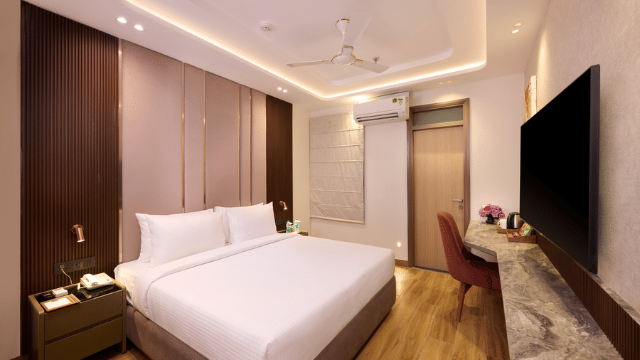 A bedroom in Tulip Inn Gurugram showing a wall-mounted flat-screen television across from a double bed with a high, padded headboard.