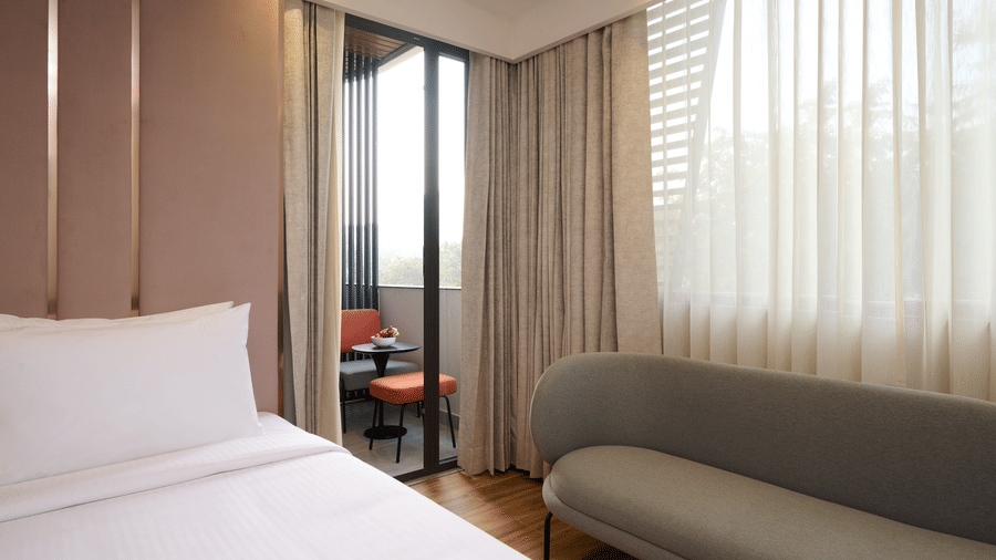 Guest room at Tulip Inn Gurugram with a plush bed in the foreground and a curved sofa placed near large windows with sheer curtains.