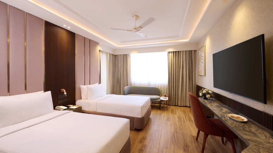 Twin bedroom at Tulip Inn Gurugram showing 2 separate beds, a long marble-topped work desk, a chair, and light-coloured curtains.