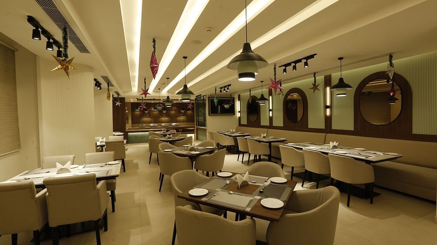  A poster of a restaurant at Tulip Inn, Zirakpur featuring several sets of tables, chairs, and sofa, lights hanging from the ceiling and mirrors attached on the wall