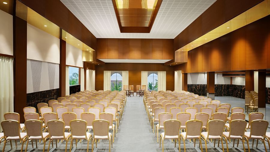 An image of a well-lit event hall with chairs arranged neatly facing a central stage, featuring modern wooden interiors, windows offering natural light, and a sleek, polished floor - Uday Backwater Resort