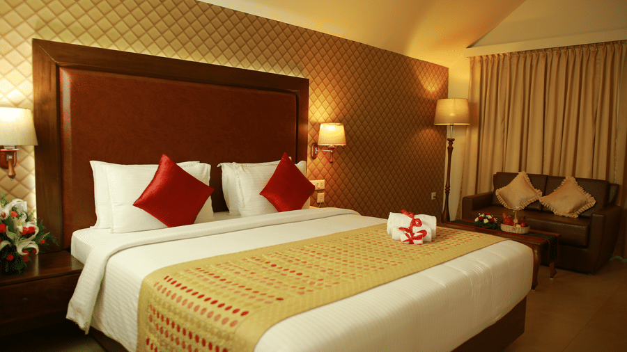 The Atrium Room at Uday Samudra Leisure Beach Hotelfurnished with a large bed featuring white linens, red pillows, and a gold and red patterned runner and includes a seating area with a dark sofa and patterned cushions