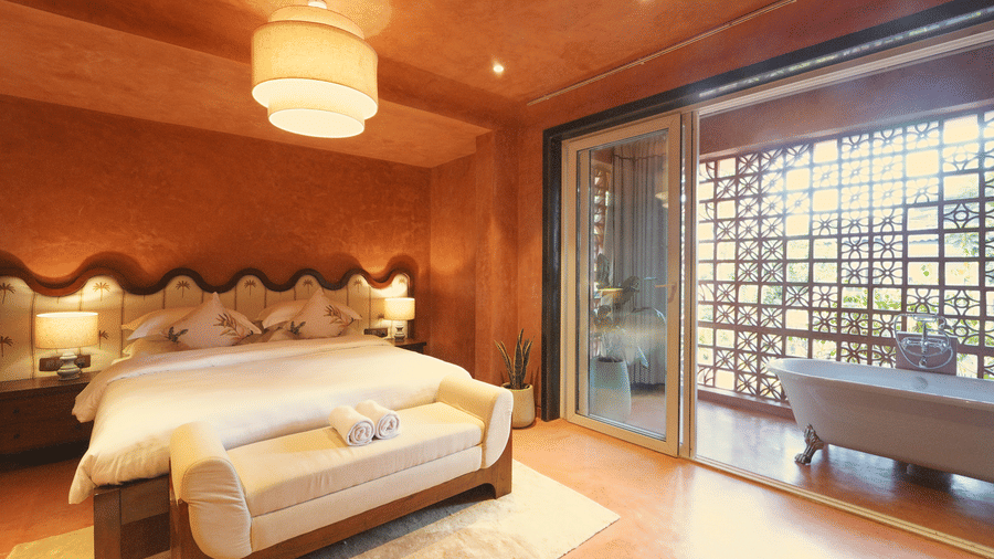 A room at Unseen Boutique Hotel Assagao Goa featuring a king bed with white bedding a wooden bench warm terracotta walls a pendant light and a glass door leading to an outdoor bathtub area.
