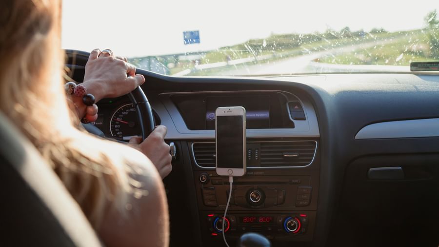 Person drives a car with a mobile phone mounted on the dashboard, view of the steering wheel, dashboard, and road ahead, related to V FIVE HOTEL.