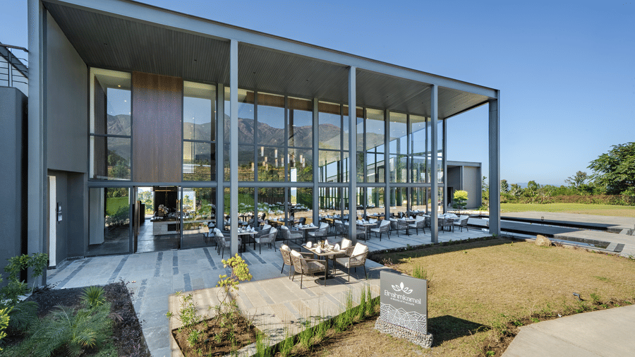 Exterior of the modern glass building, Brahmkamal Restaurant, at Vandhara Sarovar Premiere, Patkote, showing an outdoor patio area and surrounding green lawn.
