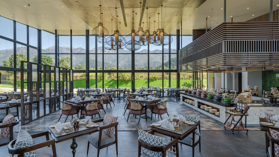 The Brahmkamal Restaurant at Vandhara Sarovar Premiere, Patkote, featuring dark wood tables, floor-to-ceiling glass walls, and hanging lantern lights.