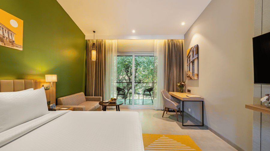 A Deluxe room at Vandhara Sarovar Premiere, Patkote, featuring a double bed, a desk area, and glass sliding doors leading to a balcony.