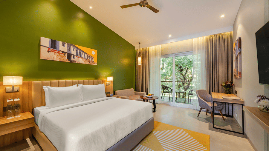 A bright Deluxe bedroom at Vandhara Sarovar Premiere, Patkote, with a ceiling fan, work desk, and views of trees through glass doors.