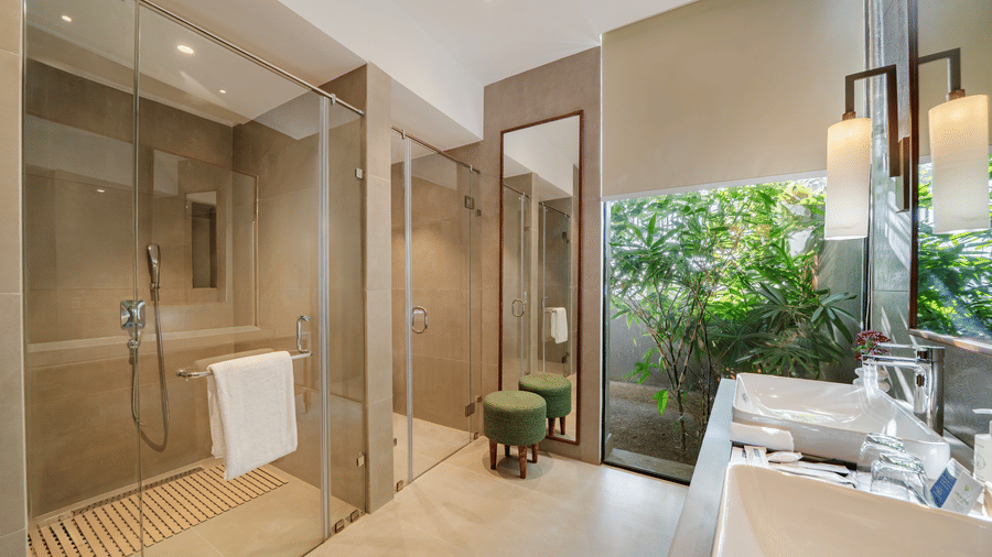 A modern bathroom at Vandhara Sarovar Premiere, Patkote, showing a walk-in glass shower, a large vanity mirror, and a window with garden views.