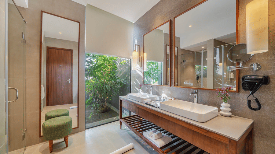 A bright bathroom at Vandhara Sarovar Premiere, Patkote, featuring a double sink vanity, large mirrors, and views of lush greenery.
