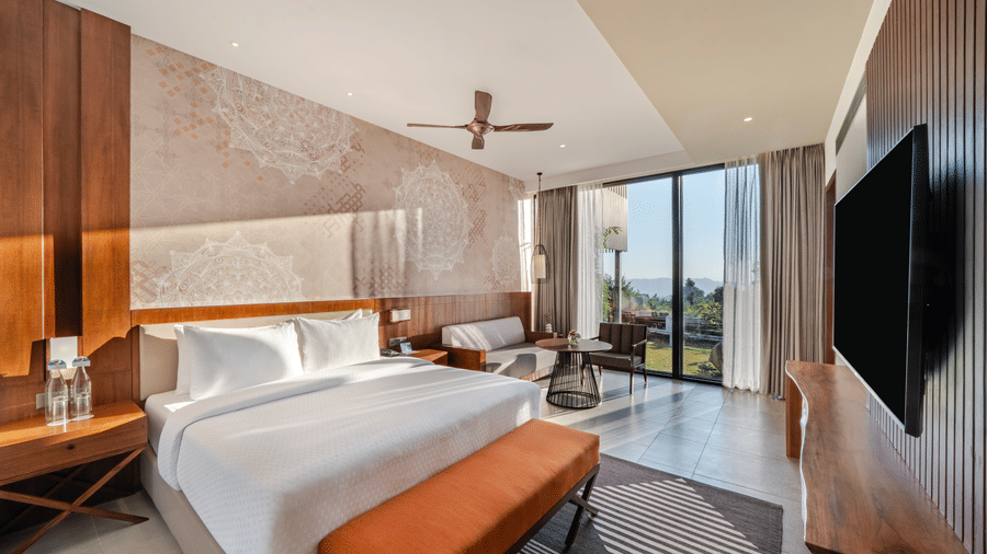 A well-lit guest room at Vandhara Sarovar Premiere, Patkote, featuring a double bed, a bench, and floor-to-ceiling windows.