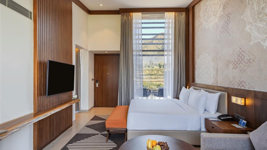 A Villa bedroom at Vandhara Sarovar Premiere, Patkote, with a king size bed, a footstool, and a large window overlooking the landscape.