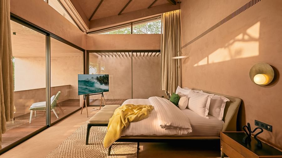 A bright bedroom at Villa Moira by V Escapes, featuring a comfortable bed, a stylish blue armchair, and large windows opening to nature.