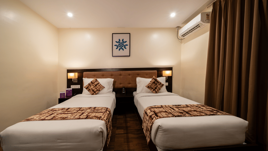 A well-lit bedroom with twin bed, small wooden side tables with lamps, ceiling lights and long curtains at VITS Select, Bengaluru.