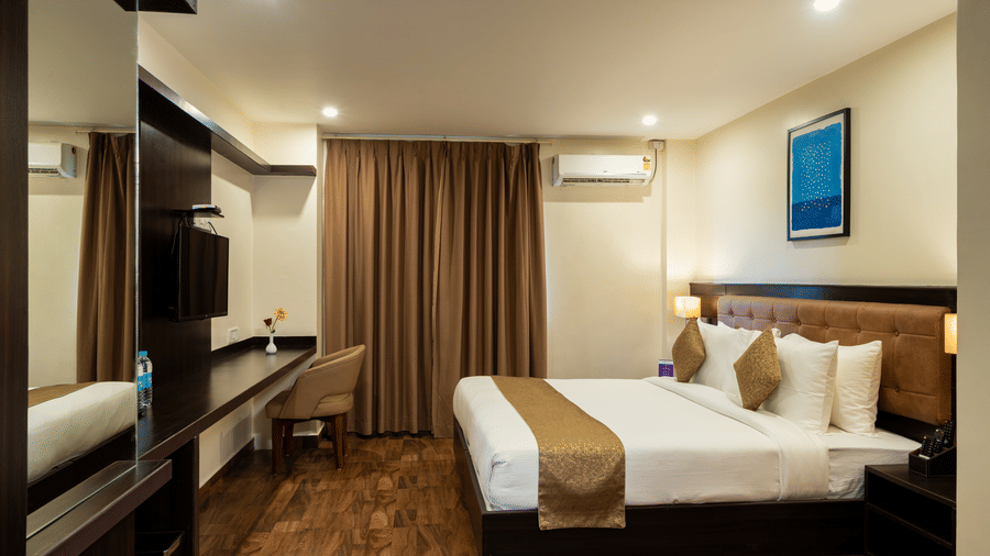 An elegant bedroom at VITS Select, Bengaluru, featuring a plush bed, rich wall paneling and curtains, a reflective mirrored surface, and a mounted air-conditioning unit for the perfect stay in Bengaluru.
