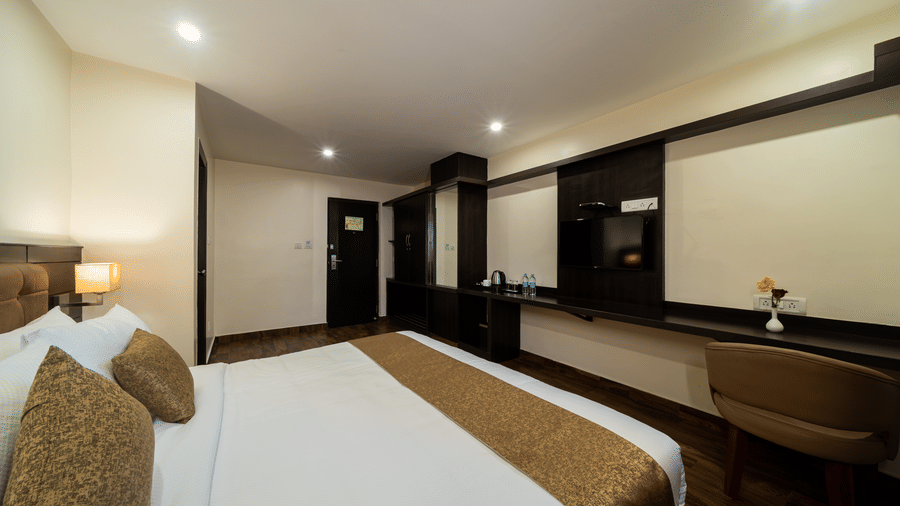 A spacious, well-organized bedroom at VITS Select, Bengaluru, showcasing a comfortably made bed, a long desk or vanity area, and a full-length mirror on a nearby cabinet.
