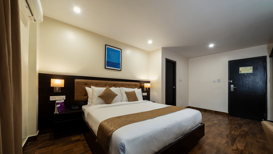 A clean, modern bedroom at VITS Select, Bengaluru, featuring a large bed, shining wooden flooring, a prominent headboard, and a closed door leading into the room.