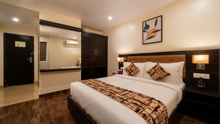 A bedroom interior at VITS Select, Bengaluru, featuring a plush bed, wooden flooring, and a bathroom entrance with mirror.
