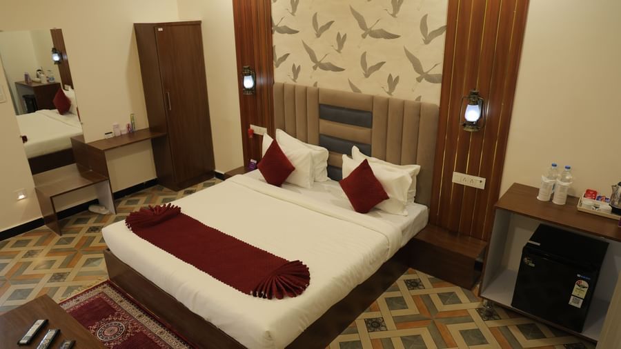 A wide shot of an Executive Room at VITS Select Diamond Resort, Sasan Gir, showing the bed with a red bed runner, a desk with a mirror, a mini-fridge, and a side table.