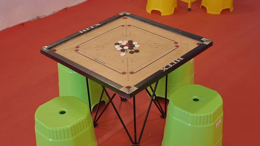 A close-up of a carrom board on a table with light green stools around it. The board has black and white pieces and a red queen.