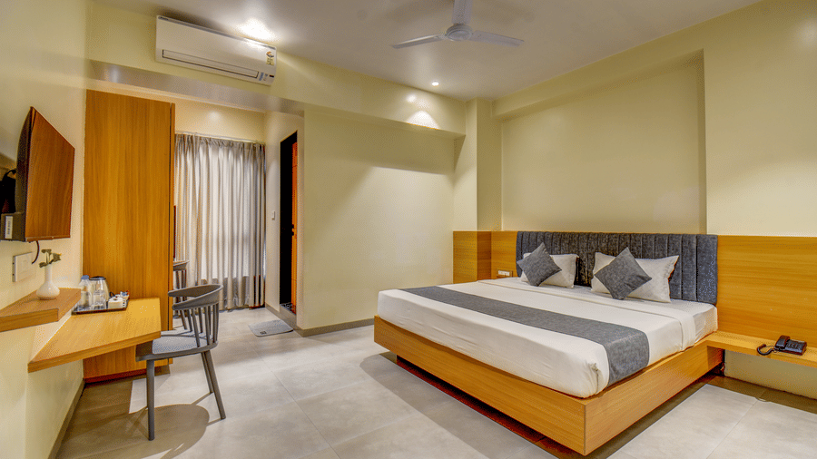 A well-lit Deluxe Room featuring a plush bed facing the TV on the wall below which is a work desk near the wardrobe