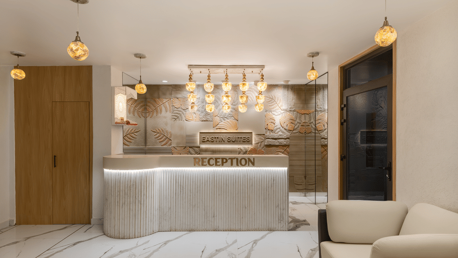 A hotel reception area featuring a curved, white reception desk with 'RECEPTION' spelled out in gold letters, decorative hanging light fixtures, and marble flooring | Voyage Eastin Suites