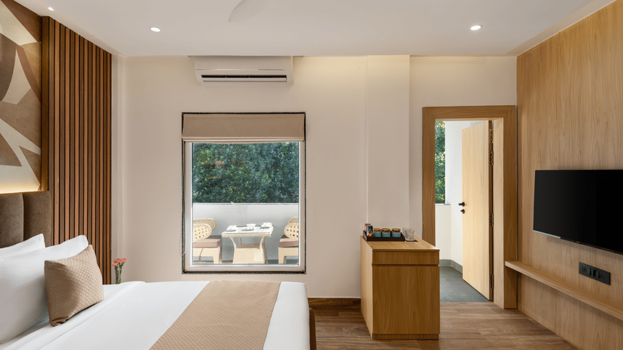A close-up view of a hotel bedroom with a bed against a feature wall, a television mounted on the wood panelling, and a glass door opening to a small outdoor area on the right | Voyage Eastin Suites