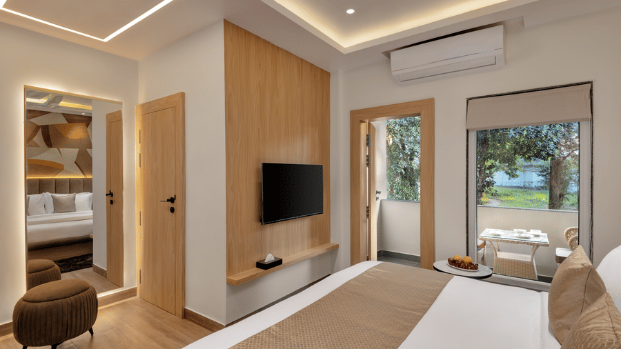 A hotel room with a large double bed, a flat-screen television on the wall, light wood panelling, and an open glass door leading to a balcony on the right | Voyage Eastin Suite