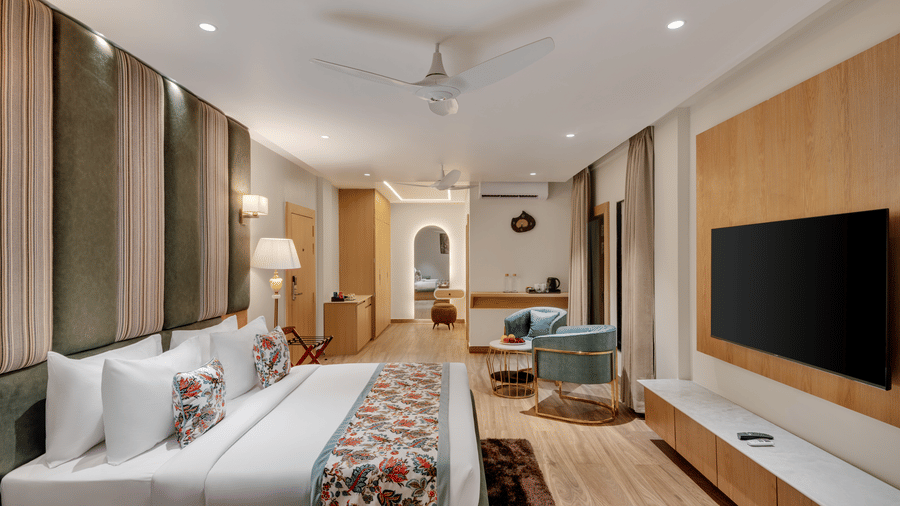 A hotel suite with a large bed in the foreground, a dark wood feature wall with a mounted television, a sitting area with a sofa and chairs near a window, and a decorative archway leading to another room | Voyage Eastin Suites