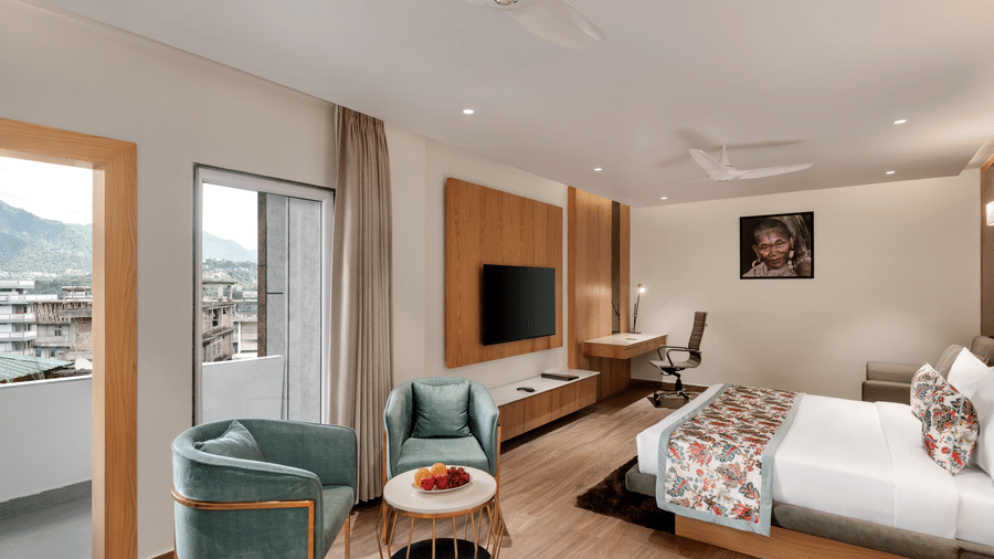 A hotel suite with a bed, an armchair and low table near the balcony doors, light wood panelling, and a view of mountains or hills outside | Voyage Eastin Suites