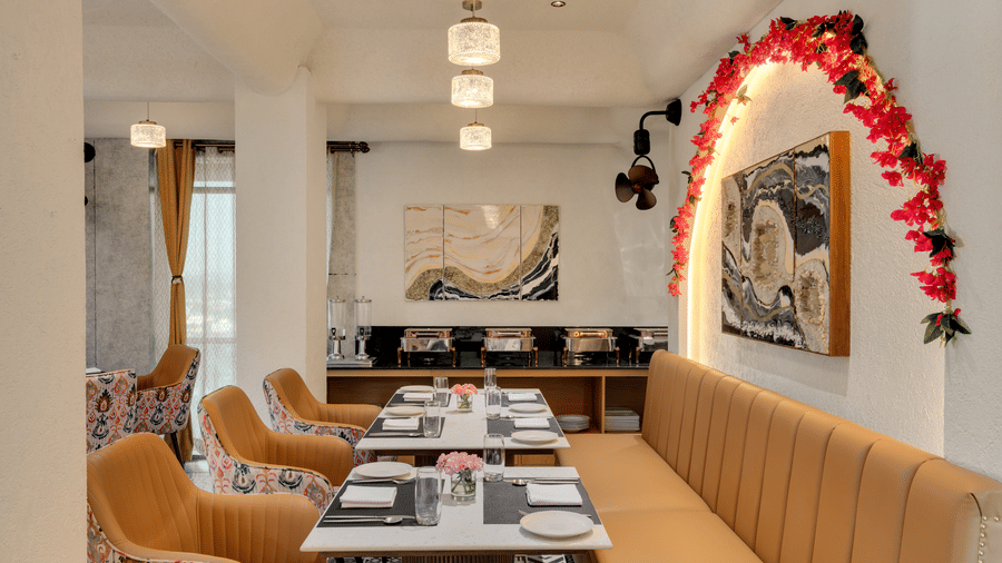 A brightly lit hotel dining area with long upholstered banquette seating against the wall, individual dining tables, and a decorative floral arch over the wall art | Voyage Eastin Suites
